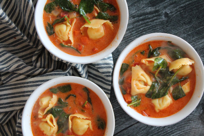 Three bowls of sun dried tomato tortellini soup on a wood surface
